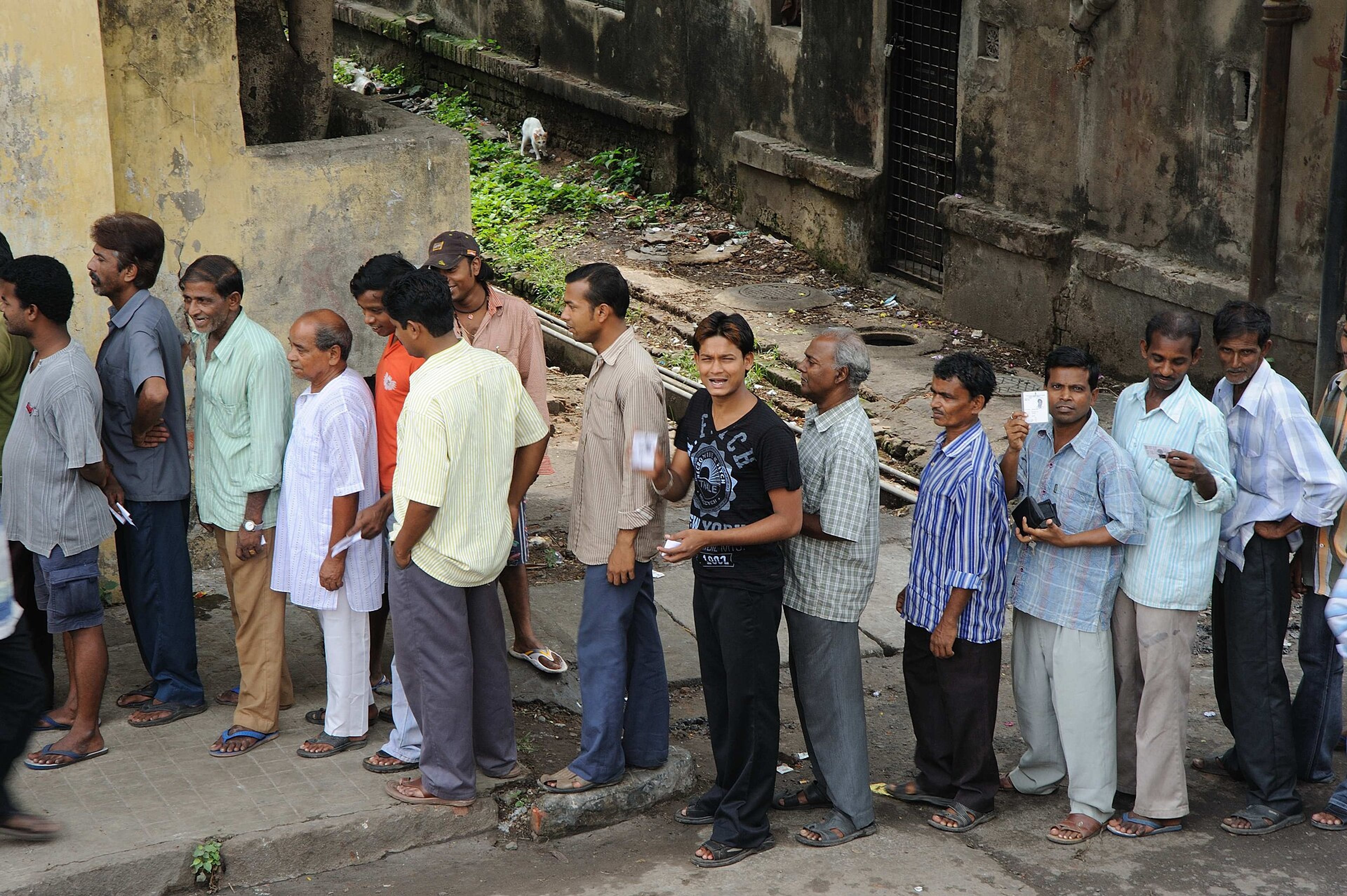 Men in casual clothes queue along a street beside an old concrete building, some holding small cards.