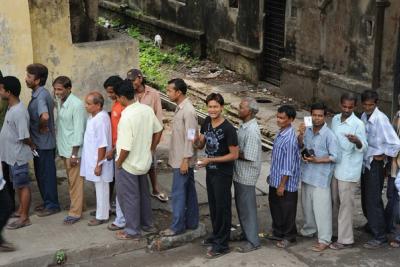 Men in casual clothes queue along a street beside an old concrete building, some holding small cards.
