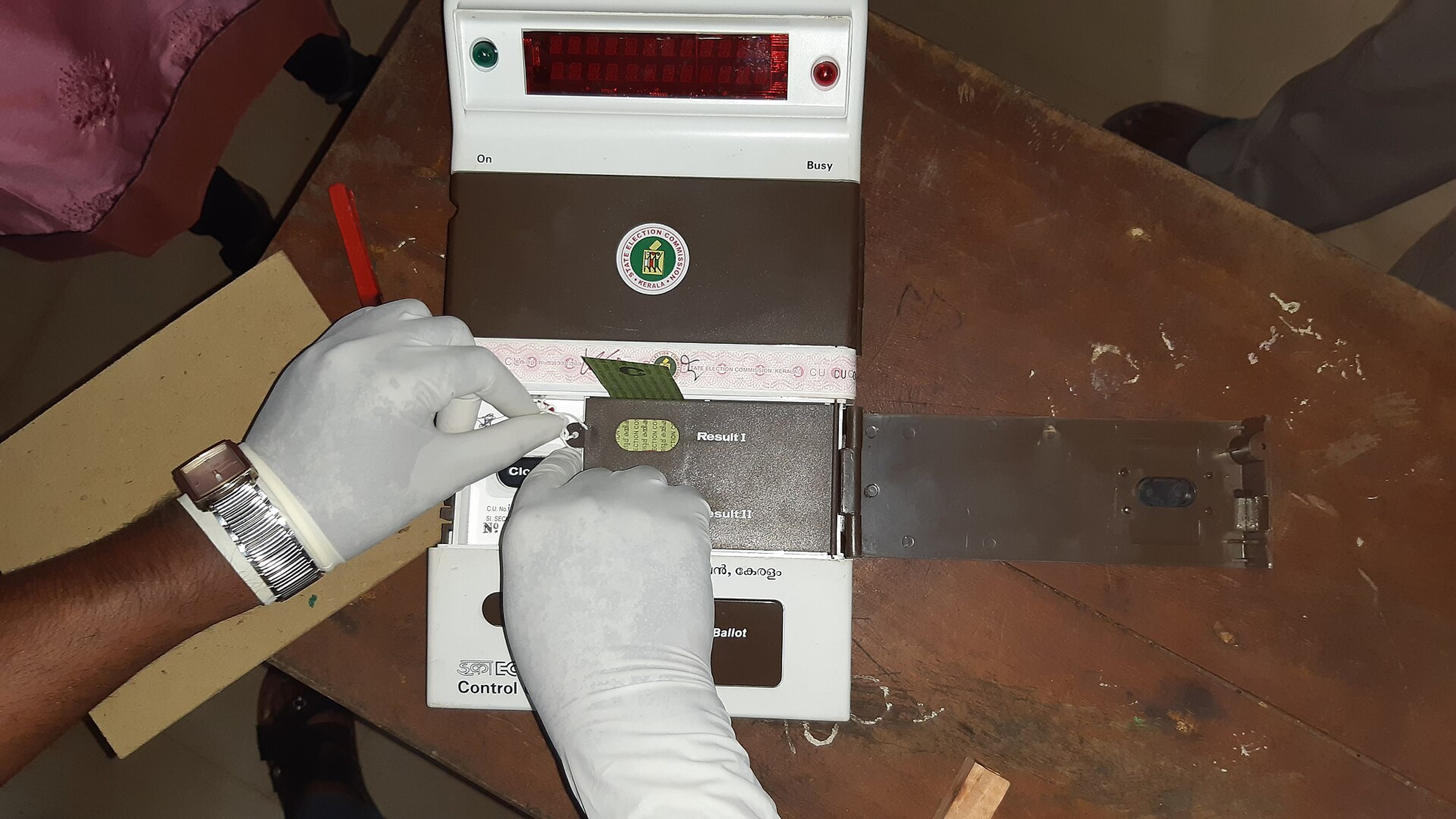 Gloved hands working on the control unit of an electronic voting machine on a wooden table.