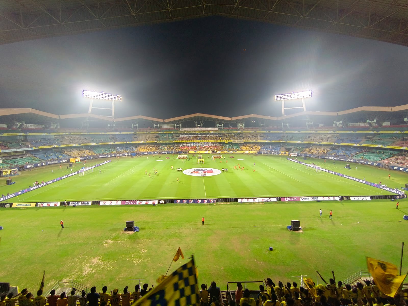 Floodlit football stadium at night with players on the pitch, Indian Super League boards and fans waving yellow flags