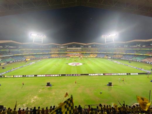 Floodlit football stadium at night with players on the pitch, Indian Super League boards and fans waving yellow flags