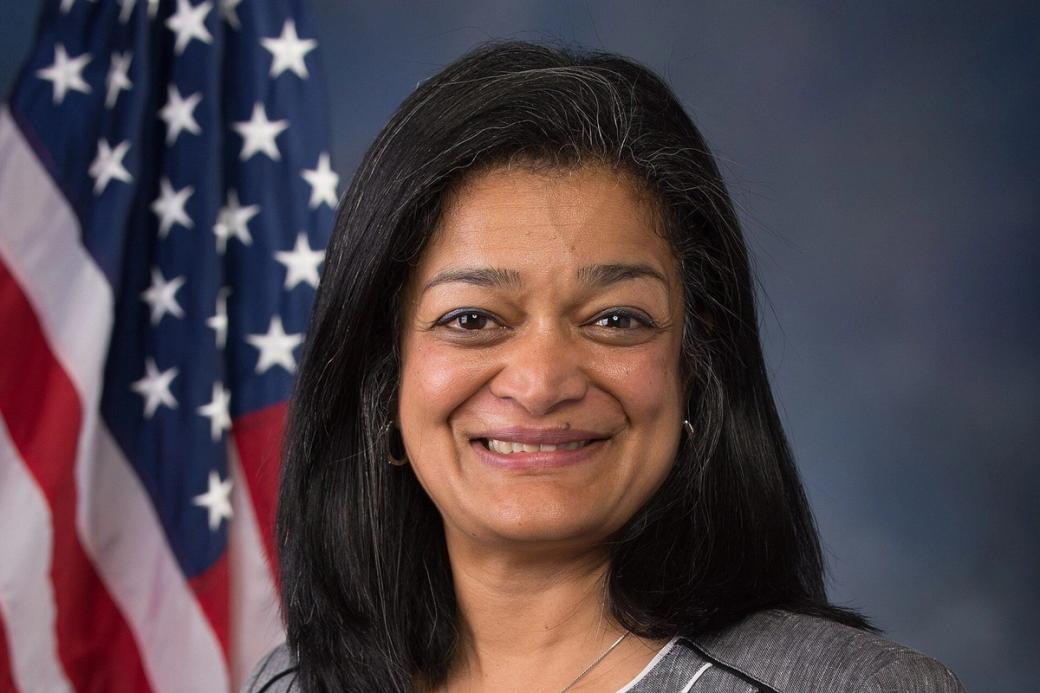 Smiling woman with shoulder-length dark hair in a grey jacket and white top, in front of an American flag and blue backdrop.