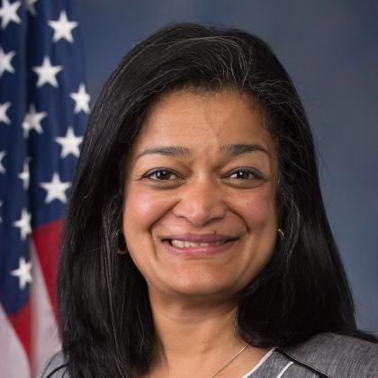 Smiling woman with shoulder-length dark hair in a grey jacket and white top, in front of an American flag and blue backdrop.