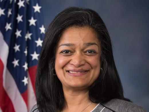 Smiling woman with shoulder-length dark hair in a grey jacket and white top, in front of an American flag and blue backdrop.