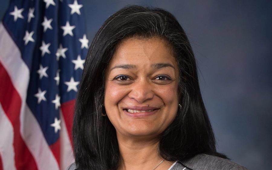 Smiling woman with shoulder-length dark hair in a grey jacket and white top, in front of an American flag and blue backdrop.