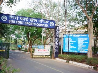 Entrance gate of the Siri Fort Sports Complex, with a blue sign, a welcome board and a facilities board under trees