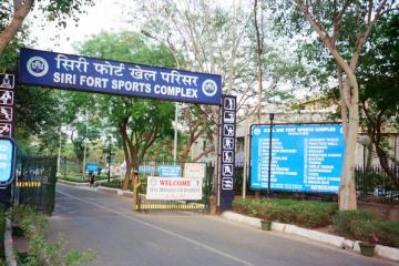 Entrance gate of the Siri Fort Sports Complex, with a blue sign, a welcome board and a facilities board under trees