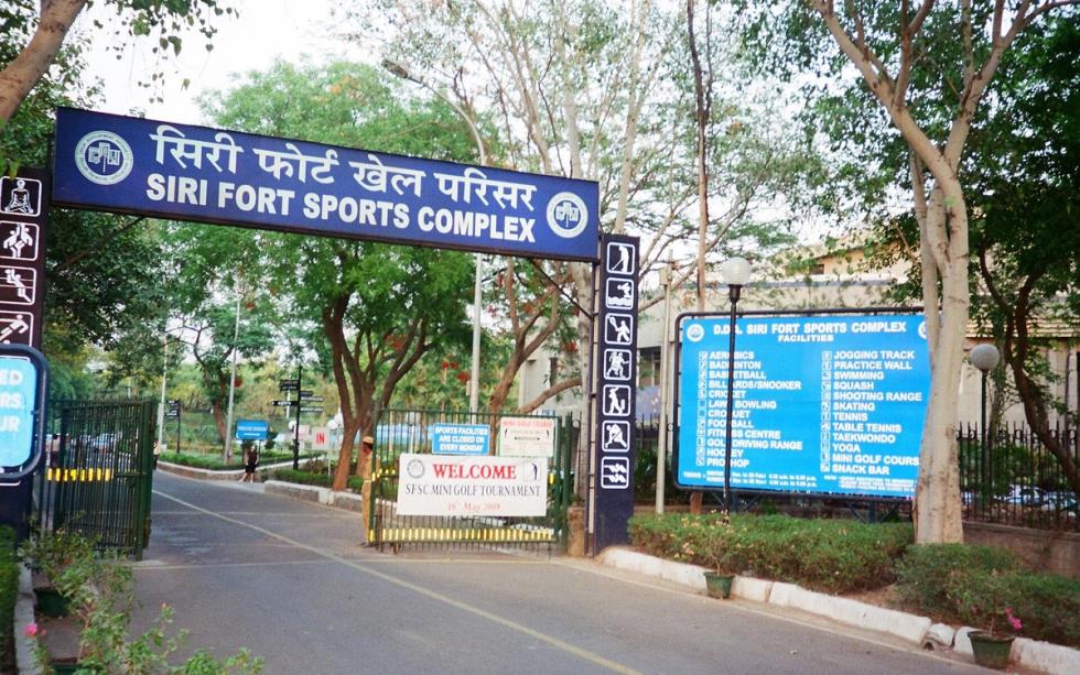 Entrance gate of the Siri Fort Sports Complex, with a blue sign, a welcome board and a facilities board under trees
