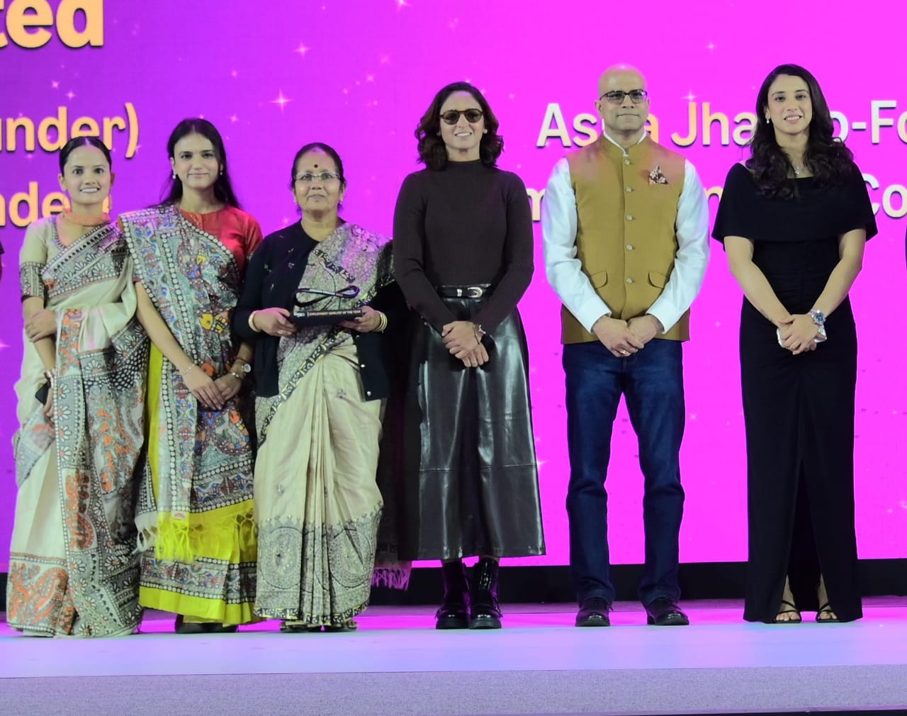 Six people pose on a stage with a magenta backdrop, three in Madhubani-print saris, one woman holding an award