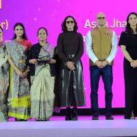 Six people pose on a stage with a magenta backdrop, three in Madhubani-print saris, one woman holding an award