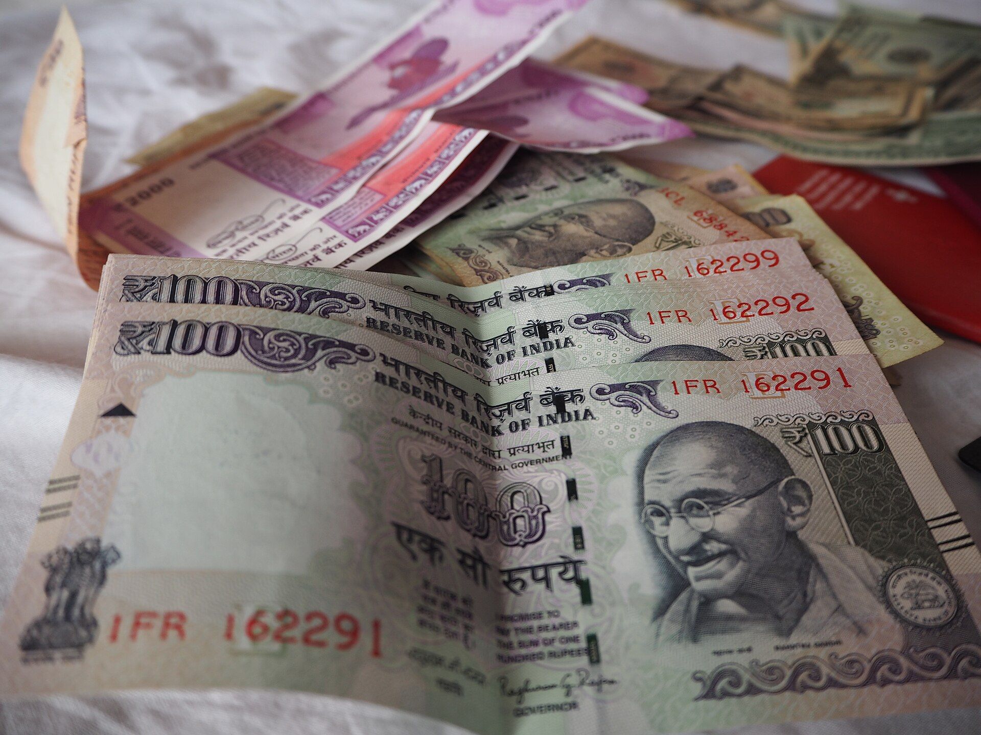 Stacked Indian ₹100 notes in the foreground, with ₹2000 notes and other currency behind them on a white cloth.