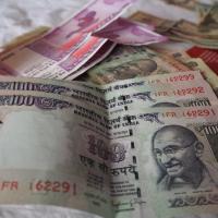 Stacked Indian ₹100 notes in the foreground, with ₹2000 notes and other currency behind them on a white cloth.