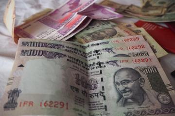Stacked Indian ₹100 notes in the foreground, with ₹2000 notes and other currency behind them on a white cloth.
