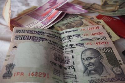 Stacked Indian ₹100 notes in the foreground, with ₹2000 notes and other currency behind them on a white cloth.