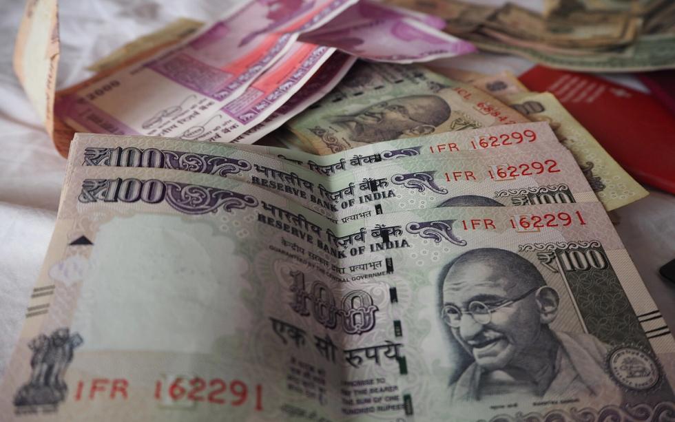 Stacked Indian ₹100 notes in the foreground, with ₹2000 notes and other currency behind them on a white cloth.