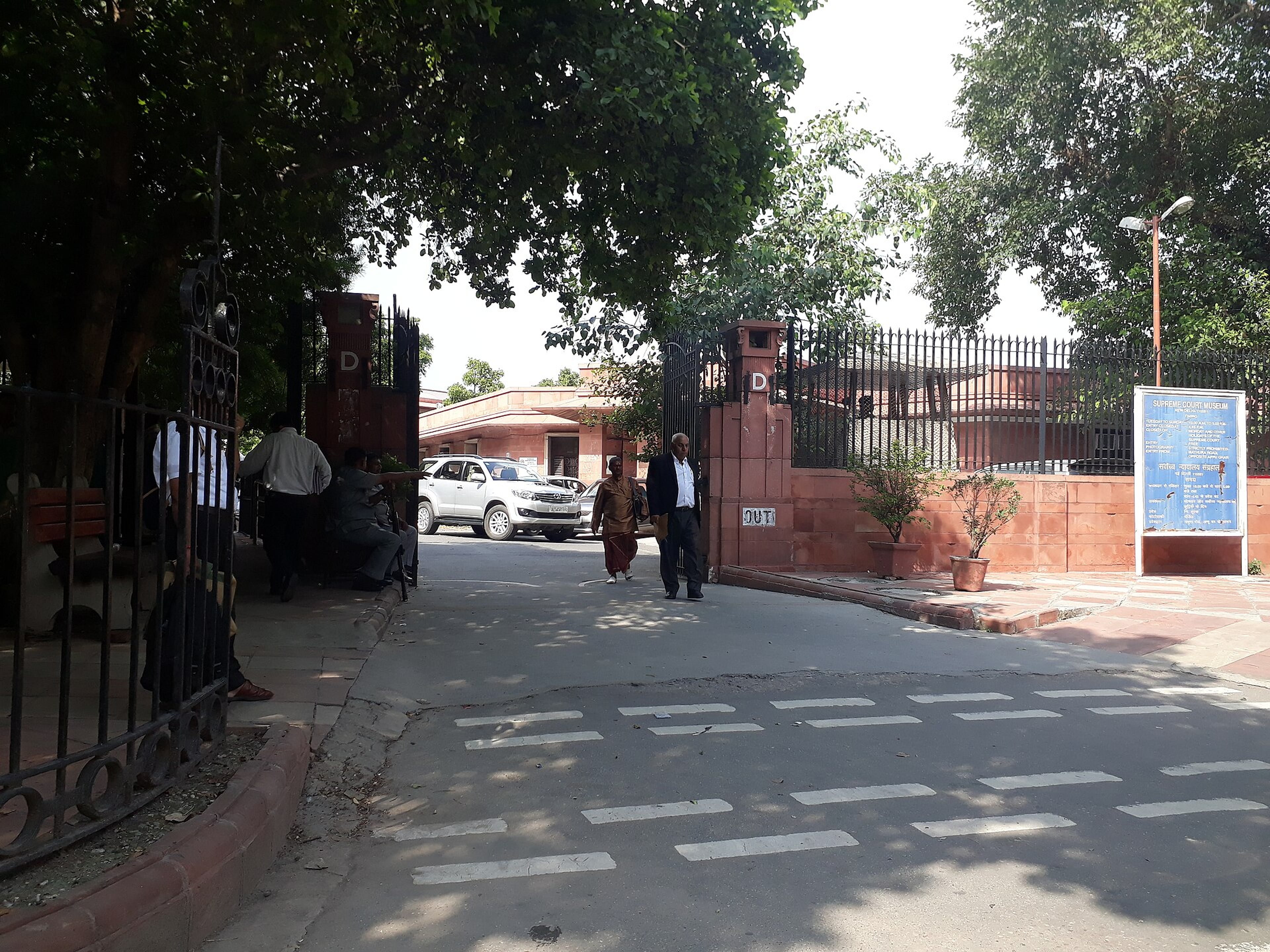 Entrance gate of the Supreme Court of India in New Delhi, with a parked car and people walking past under trees.