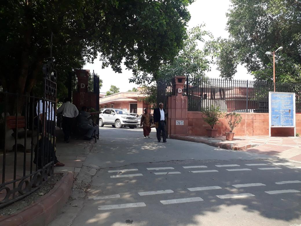 Entrance gate of the Supreme Court of India in New Delhi, with a parked car and people walking past under trees.