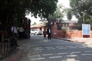 Entrance gate of the Supreme Court of India in New Delhi, with a parked car and people walking past under trees.