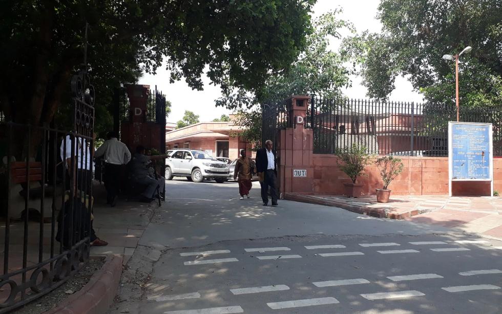 Entrance gate of the Supreme Court of India in New Delhi, with a parked car and people walking past under trees.