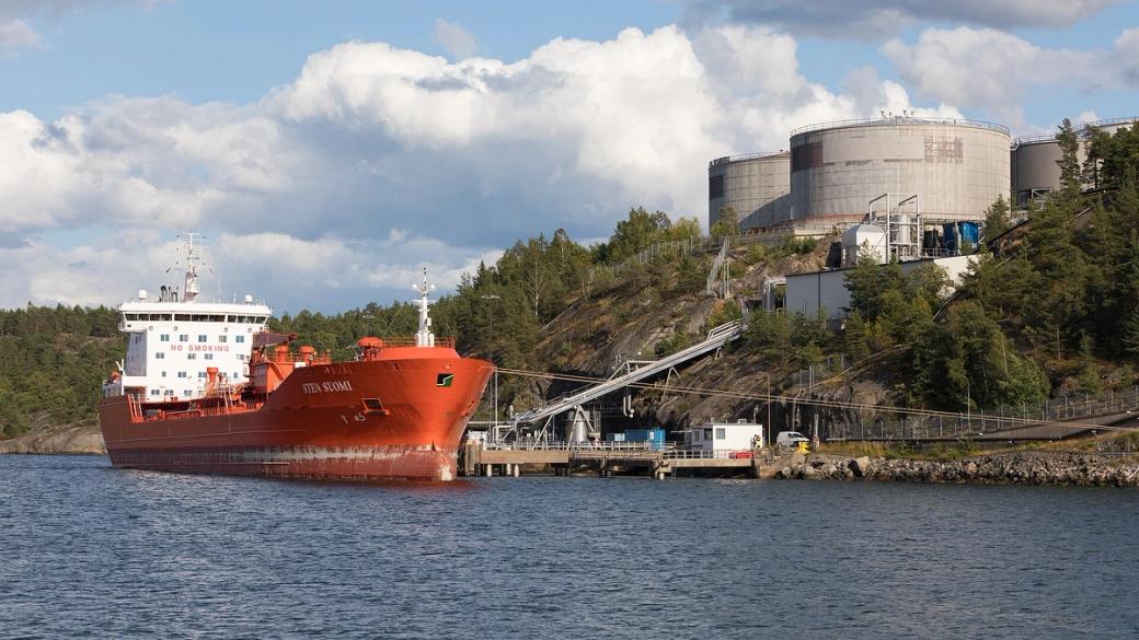 Orange oil tanker Sten Suomi moored beside a fuel depot with large storage tanks on a wooded shore.