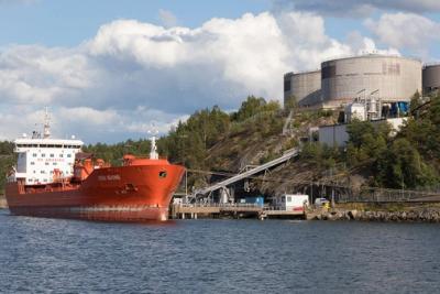 Orange oil tanker Sten Suomi moored beside a fuel depot with large storage tanks on a wooded shore.