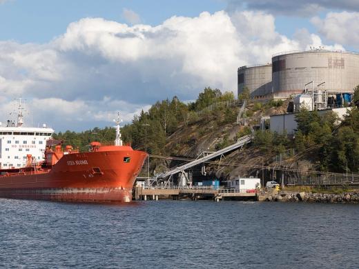 Orange oil tanker Sten Suomi moored beside a fuel depot with large storage tanks on a wooded shore.