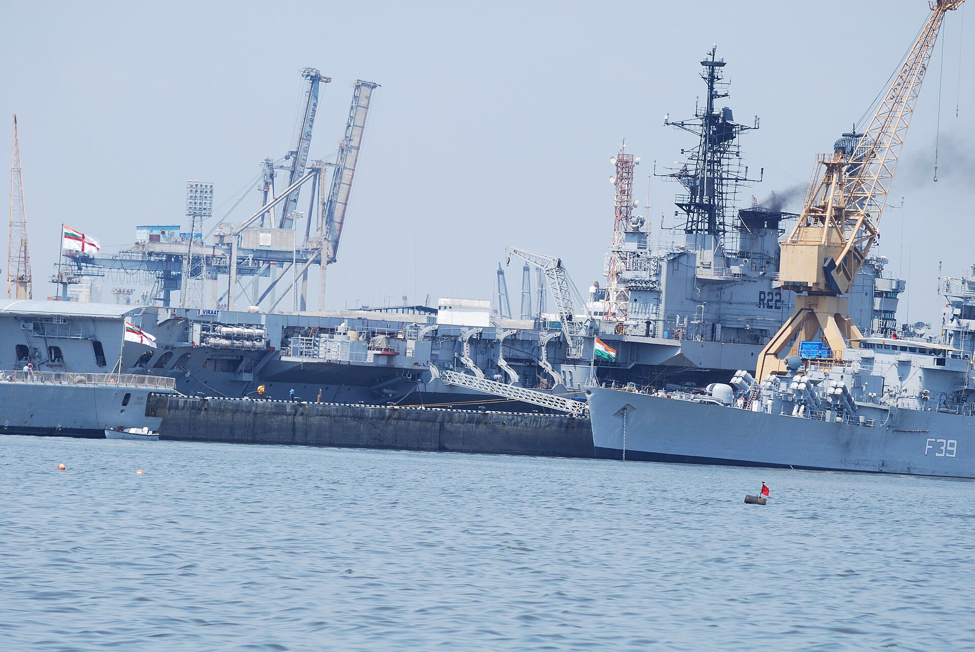 Indian Navy warships moored at a harbour quay beside port cranes, under a hazy sky.