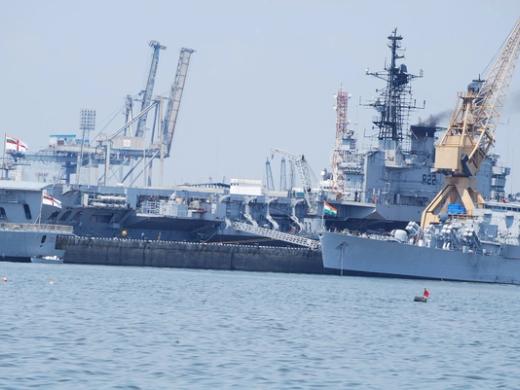 Indian Navy warships moored at a harbour quay beside port cranes, under a hazy sky.