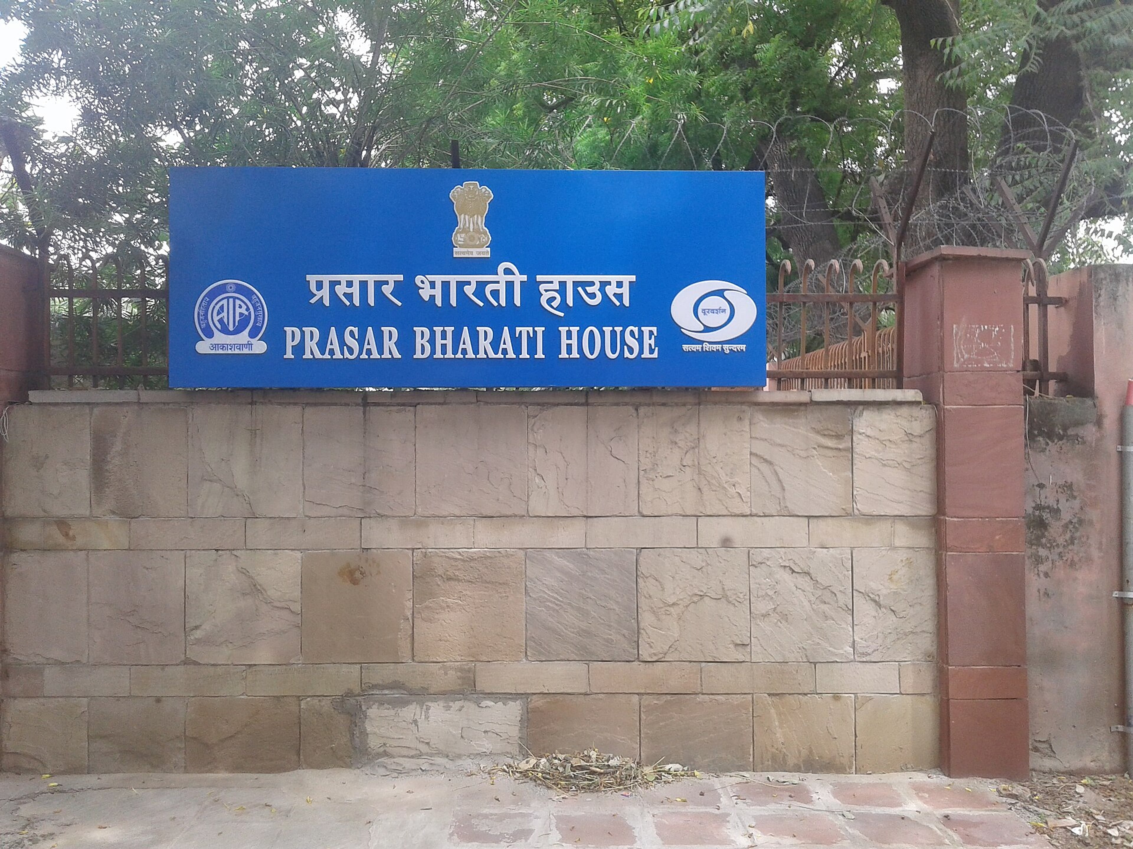 Blue sign reading Prasar Bharati House in Hindi and English on a stone wall in front of trees