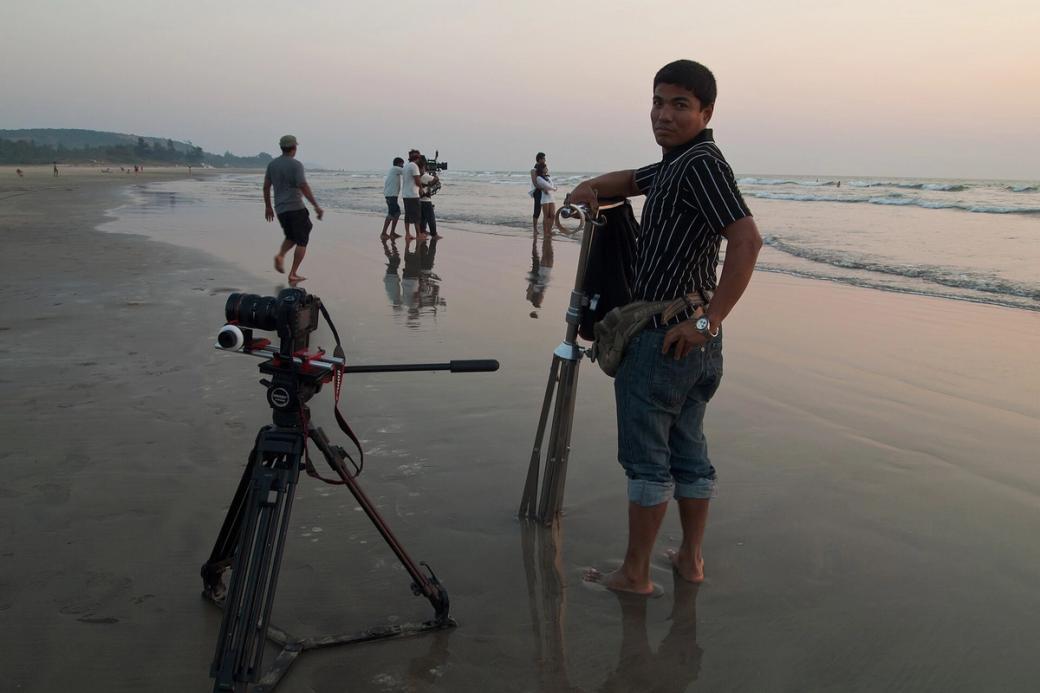 Camera on a tripod on a wet beach at dusk, with crew members filming and a man beside a second tripod.