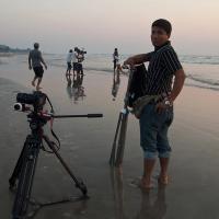 Camera on a tripod on a wet beach at dusk, with crew members filming and a man beside a second tripod.