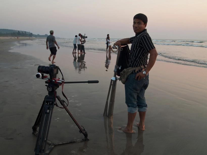 Camera on a tripod on a wet beach at dusk, with crew members filming and a man beside a second tripod.
