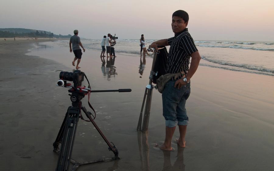 Camera on a tripod on a wet beach at dusk, with crew members filming and a man beside a second tripod.