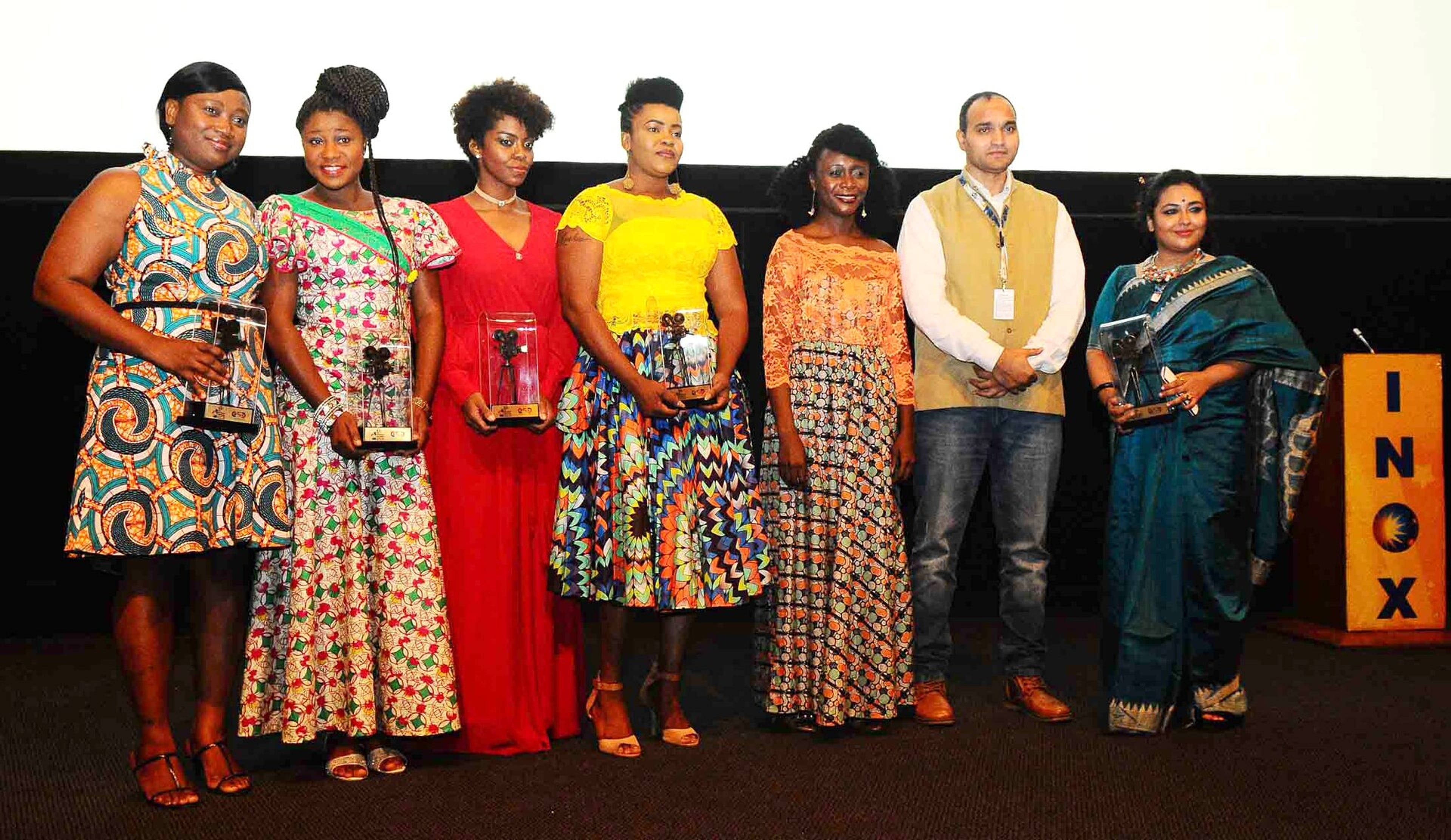 Seven people in colourful outfits hold glass trophies on a stage in front of a cinema screen.