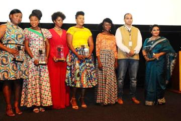 Seven people in colourful outfits hold glass trophies on a stage in front of a cinema screen.