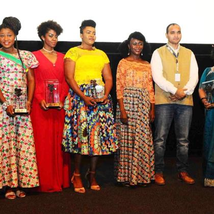 Seven people in colourful outfits hold glass trophies on a stage in front of a cinema screen.