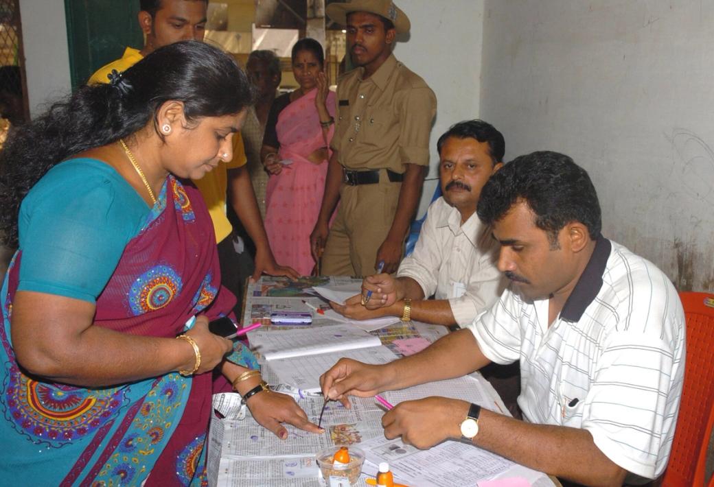 Polling officer applies indelible ink to a woman voter's finger at a booth, with officials and a policeman at a table behind