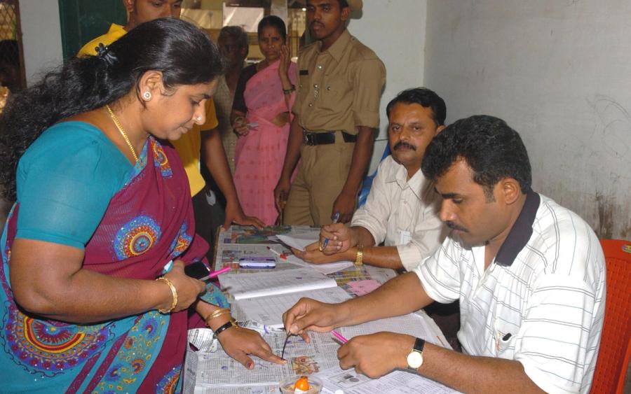 Polling officer applies indelible ink to a woman voter's finger at a booth, with officials and a policeman at a table behind