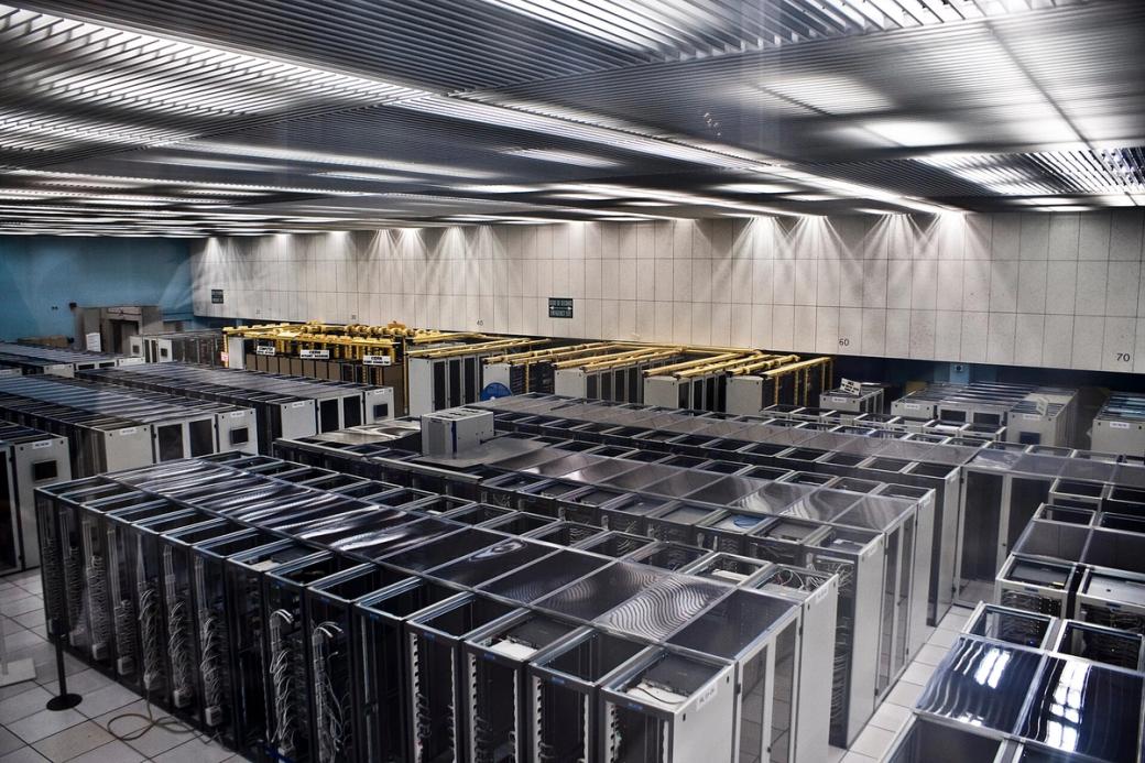 Rows of server racks in a large data centre room with overhead lighting and white floor tiles.