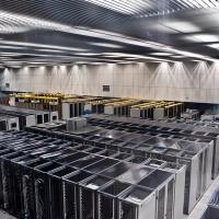 Rows of server racks in a large data centre room with overhead lighting and white floor tiles.