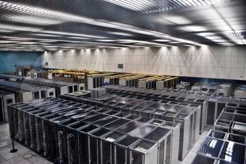 Rows of server racks in a large data centre room with overhead lighting and white floor tiles.