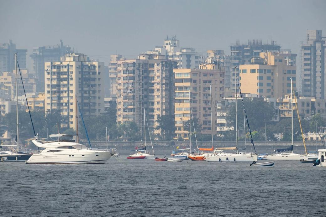 Sailboats and motor yachts moored on hazy grey water in front of mid-rise buildings and trees along a shoreline.