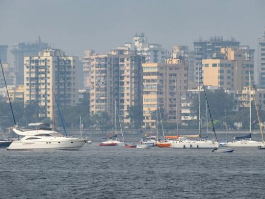Sailboats and motor yachts moored on hazy grey water in front of mid-rise buildings and trees along a shoreline.