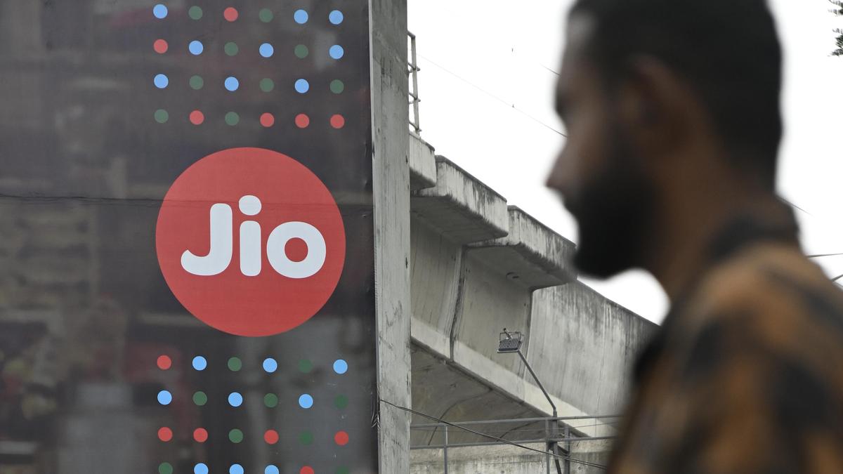 A passer-by, out of focus, walks past a red circular Jio sign on a hoarding beside a concrete flyover