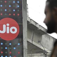 A passer-by, out of focus, walks past a red circular Jio sign on a hoarding beside a concrete flyover