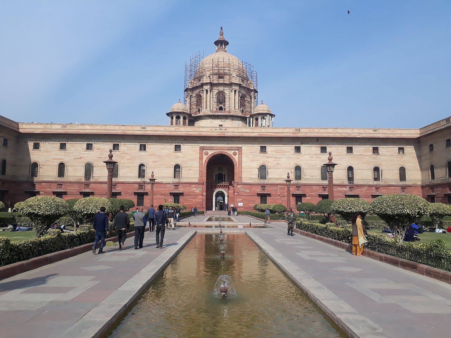 Sandstone North Block with a central dome and arched entrance, seen across a fountain pool with people on the paved paths