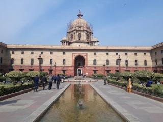 Sandstone North Block with a central dome and arched entrance, seen across a fountain pool with people on the paved paths