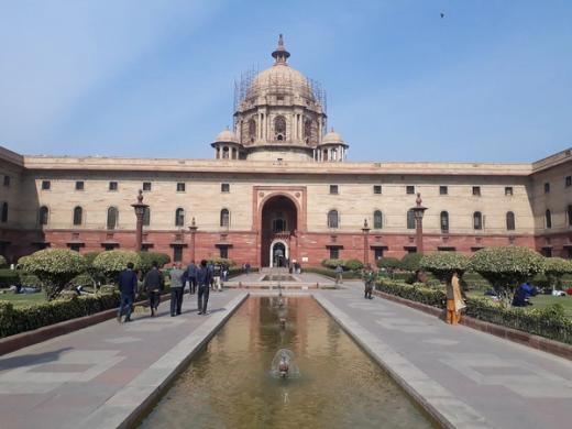 Sandstone North Block with a central dome and arched entrance, seen across a fountain pool with people on the paved paths
