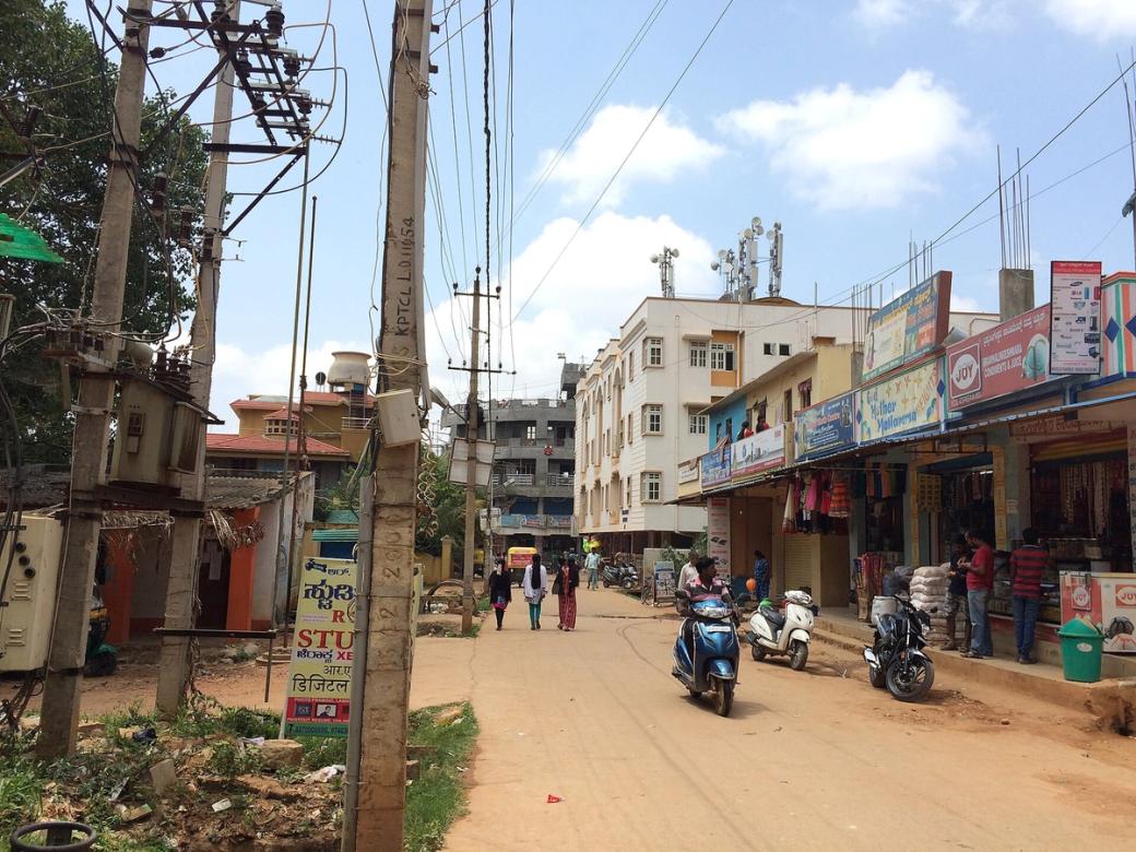 Busy street lined with shops, motorbikes and pedestrians in Bengaluru under a partly cloudy sky.