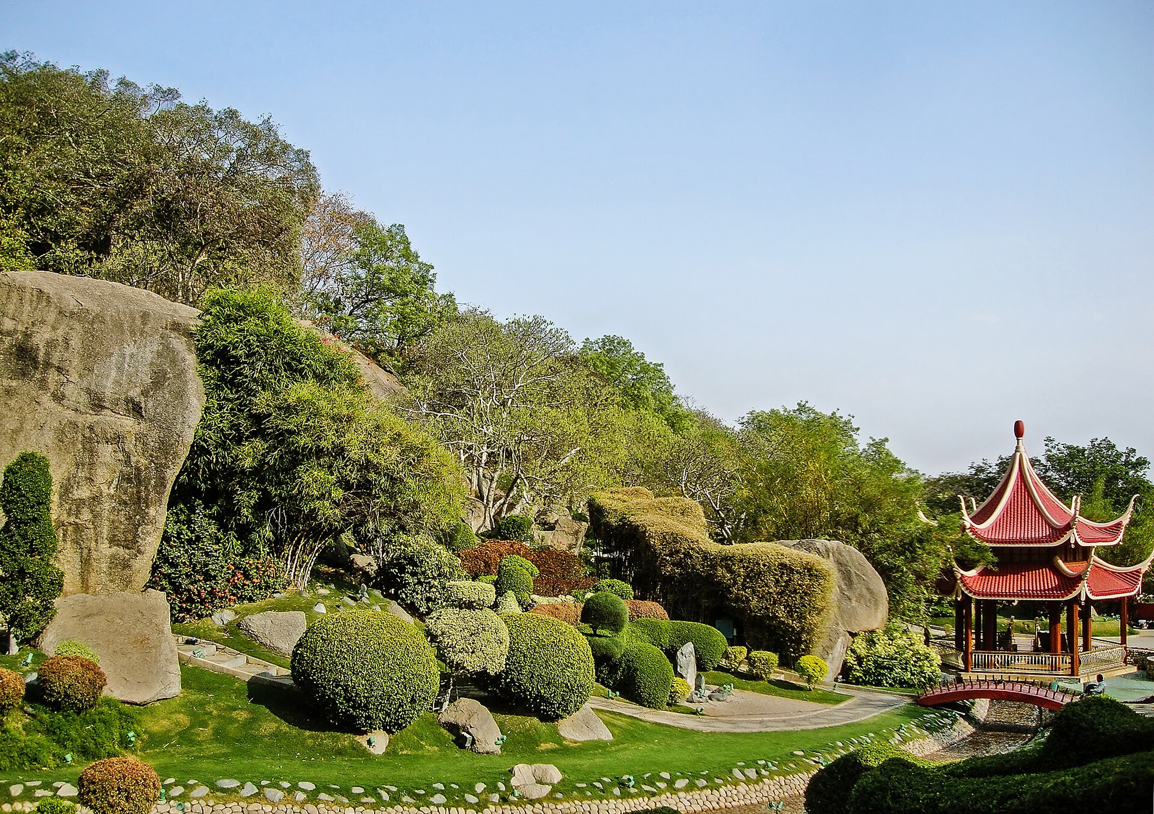 Landscaped garden with large boulders, clipped shrubs and a red pagoda beside a stone path under a clear sky